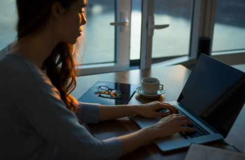 Chica escribiendo en computadora con libreta, taza y anteojos sobre la mesa